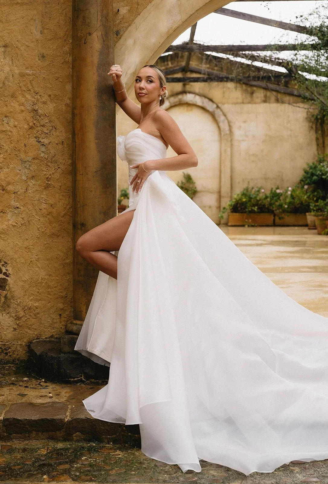 A woman in a strapless white wedding dress with a long train, standing by an archway, with one leg slightly raised.