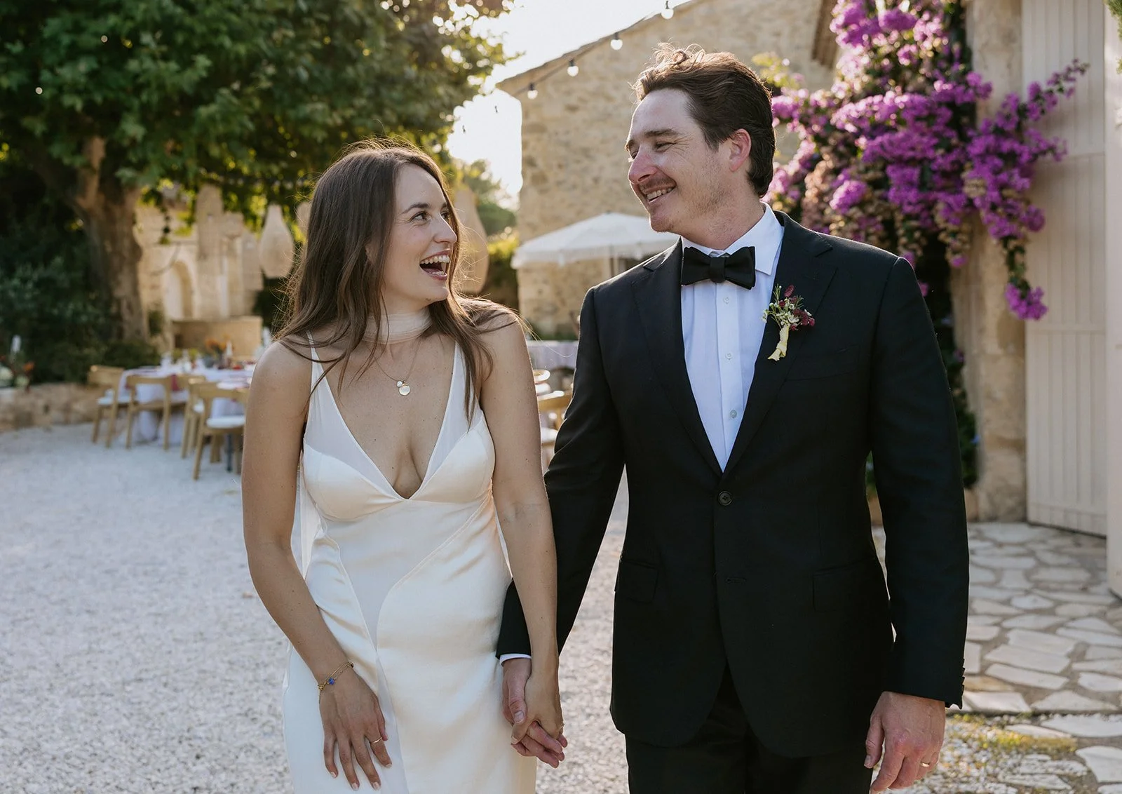 Brontë McVeigh wedding photography: A bride and groom walking outdoors and smiling at each other, holding hands during their wedding celebration near a stone building with purple flowers.