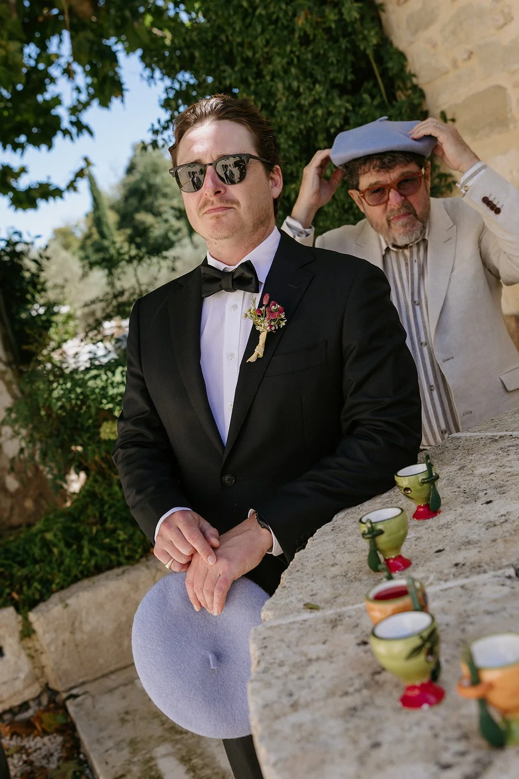 A man in a tuxedo with sunglasses and a boutonniere sitting outdoors, holding a hat. Behind him, a man in a light-colored suit and sunglasses adjusts a blue hat. There are colorful cups on a stone surface in the foreground, and trees and a wall in th