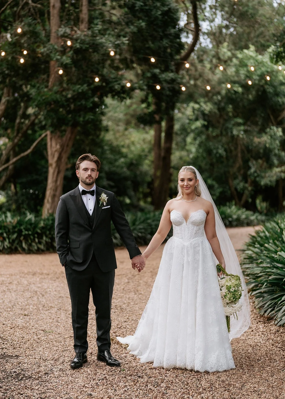 A bride and groom standing outdoors on a gravel path, holding hands, surrounded by trees and string lights in a garden setting.