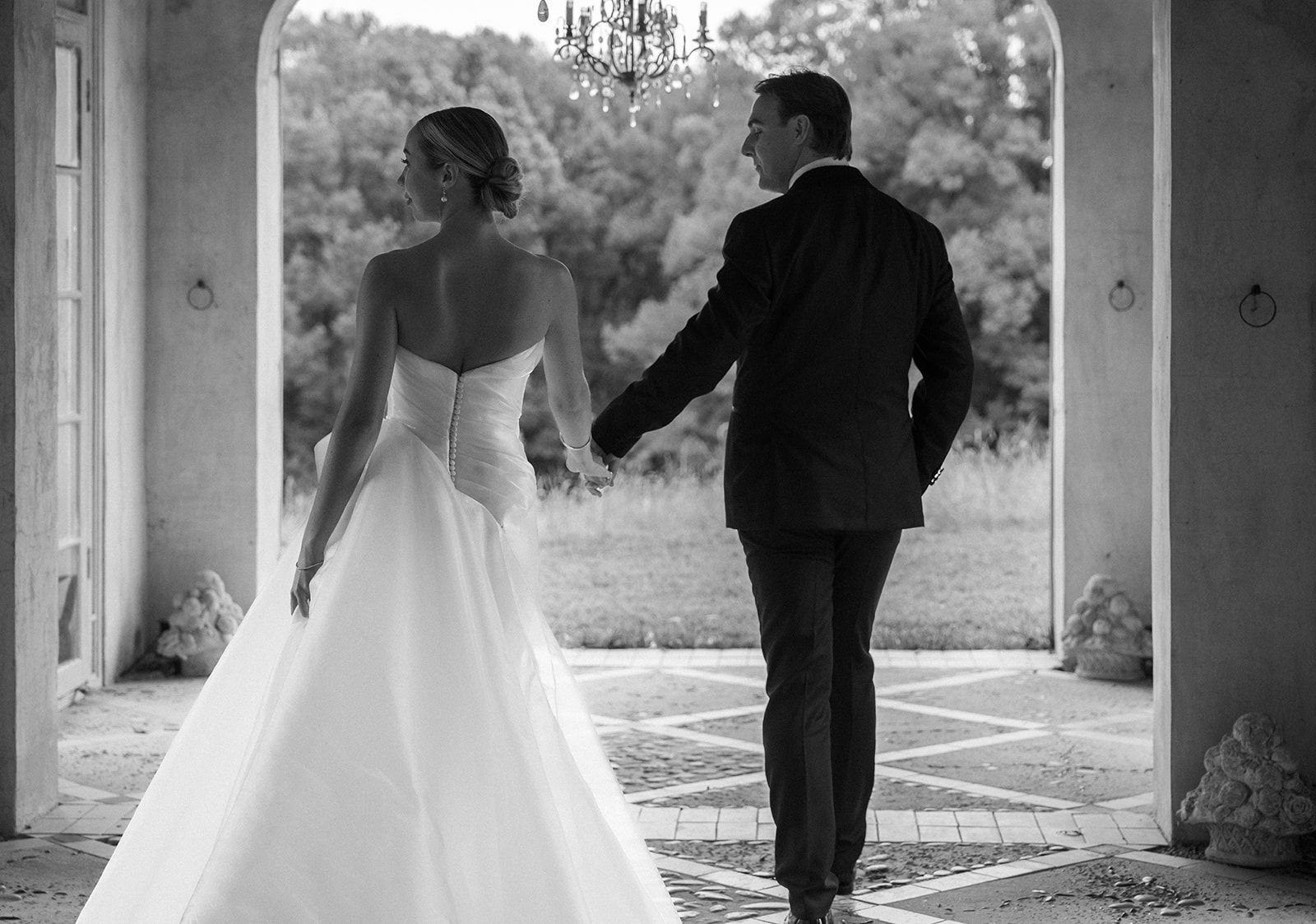 A black and white photo of a bride and groom holding hands, walking away from the camera through an archway, with a scenic outdoor background.