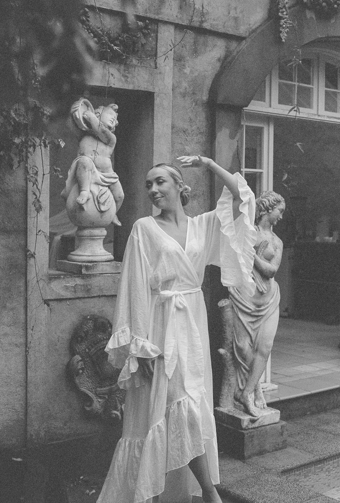 A woman in a flowing white dress poses with her hand raised above her head, standing next to classical statues and sculptures in an outdoor setting.