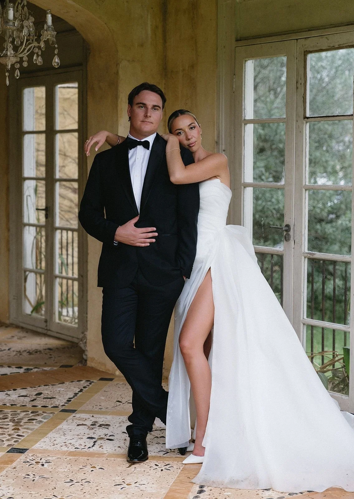 A newlywed couple in formal wedding attire posing inside a rustic house with large windows and a chandelier.