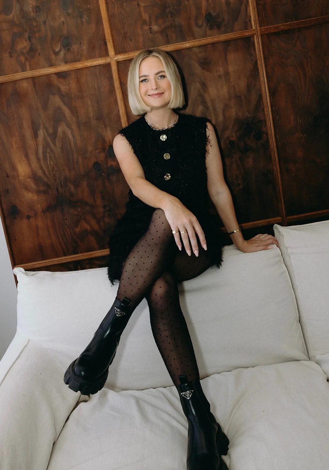 Brontë McVeigh sitting on a cream-colored couch, wearing a black sleeveless dress with three large gold buttons, patterned black tights, and chunky black boots. She has short blonde hair and is smiling at the camera against a wooden paneled wall.