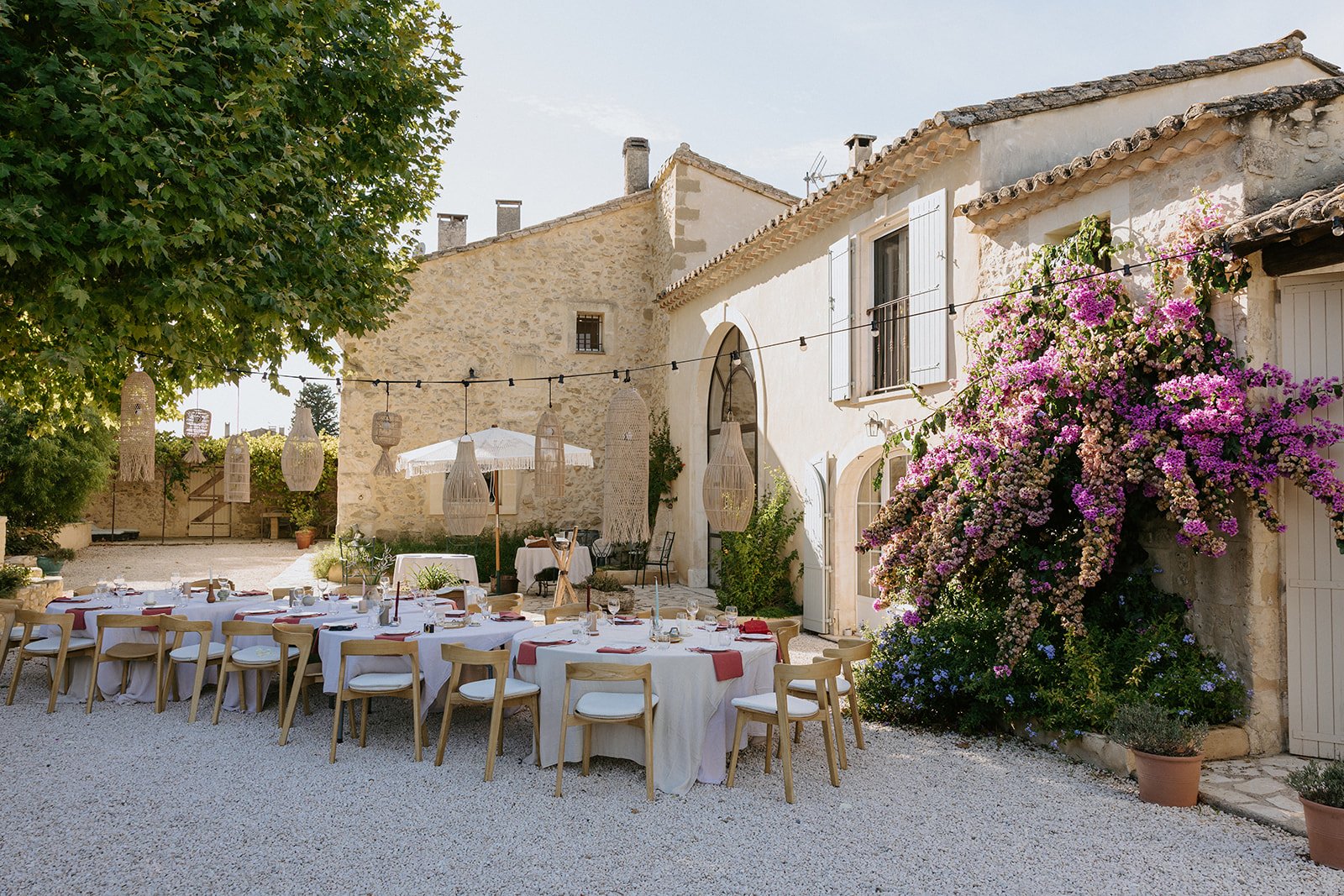 Outdoor wedding or event setup with round tables, white tablecloths, pink napkins, and chairs, set in a courtyard with stone buildings, a large tree, pink flowering bushes, and string lights hanging overhead.
