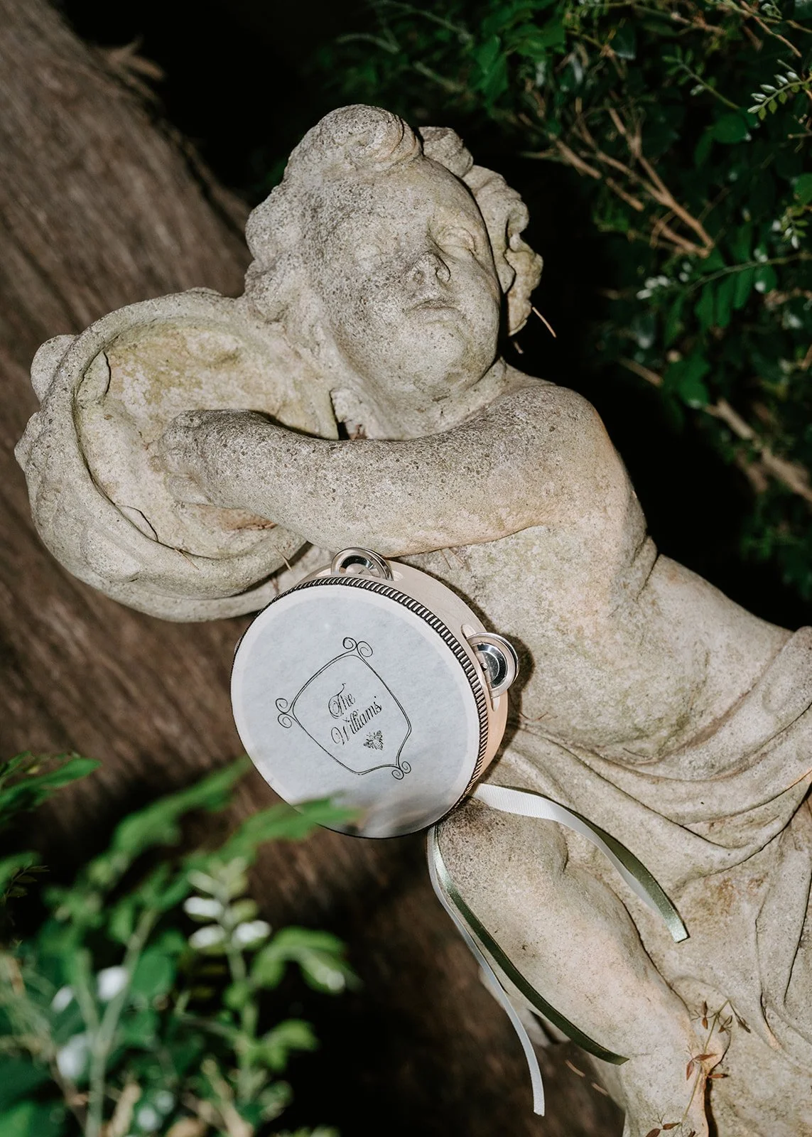 A stone sculpture of a cherub lying down with crossed arms, holding a small, round, metallic purse with the words 'The Williams' inscribed on it. The sculpture is outdoors, surrounded by greenery.