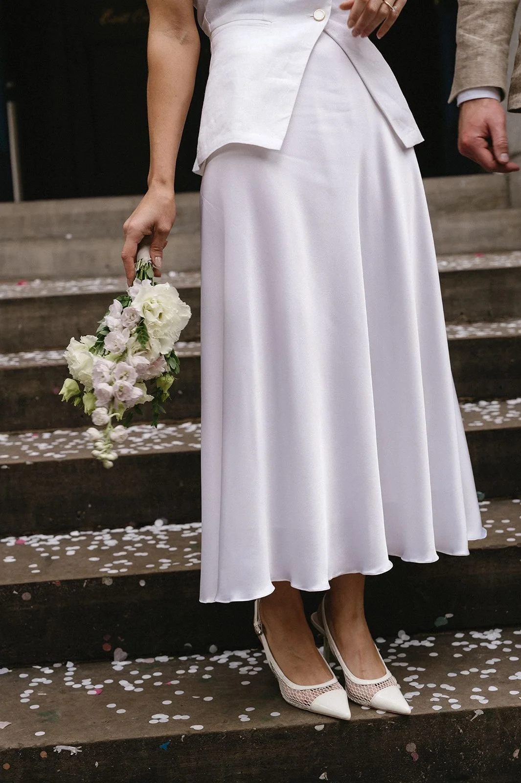 Woman in a white dress holding a bouquet of white and pink flowers, standing on steps covered with confetti, wearing white high heels.