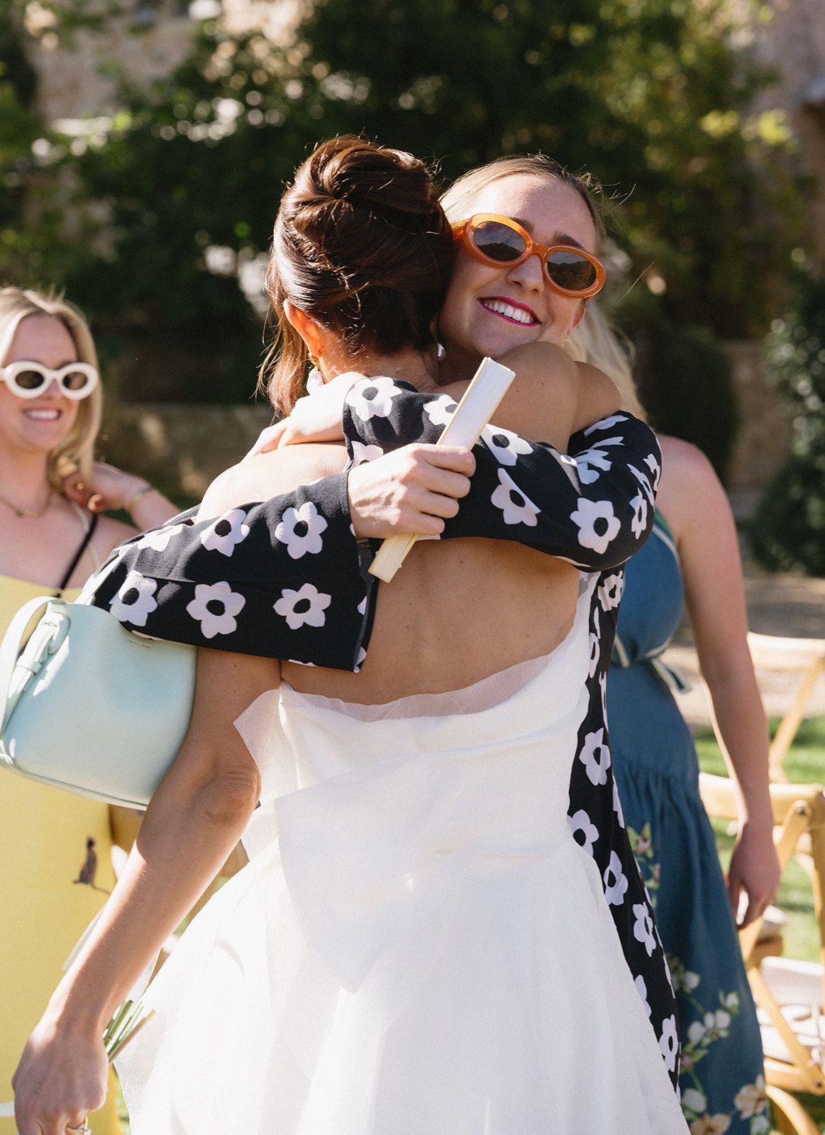 Two women warmly hugging, one smiling brightly with orange sunglasses, the other with brunette hair styled in an updo and wearing a floral black and white jacket. In the background, two other women in summer dresses, one with white sunglasses, appear
