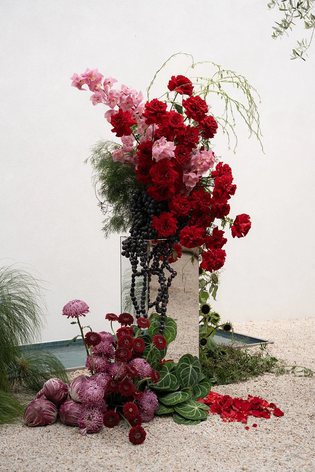 A floral arrangement with pink, red, and purple flowers, including roses, dahlias, and anthuriums, displayed on a pedestal with black beads and green foliage, set against a white background with some greenery.