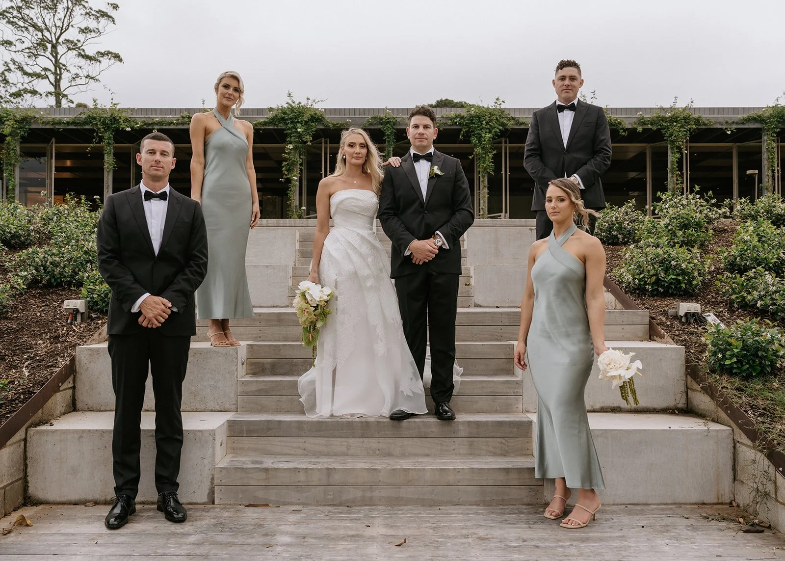Group of seven people, two women in gray dresses, three men in black tuxedos, and two women in wedding dresses, standing on steps outdoors during daytime with greenery and modern building in background.