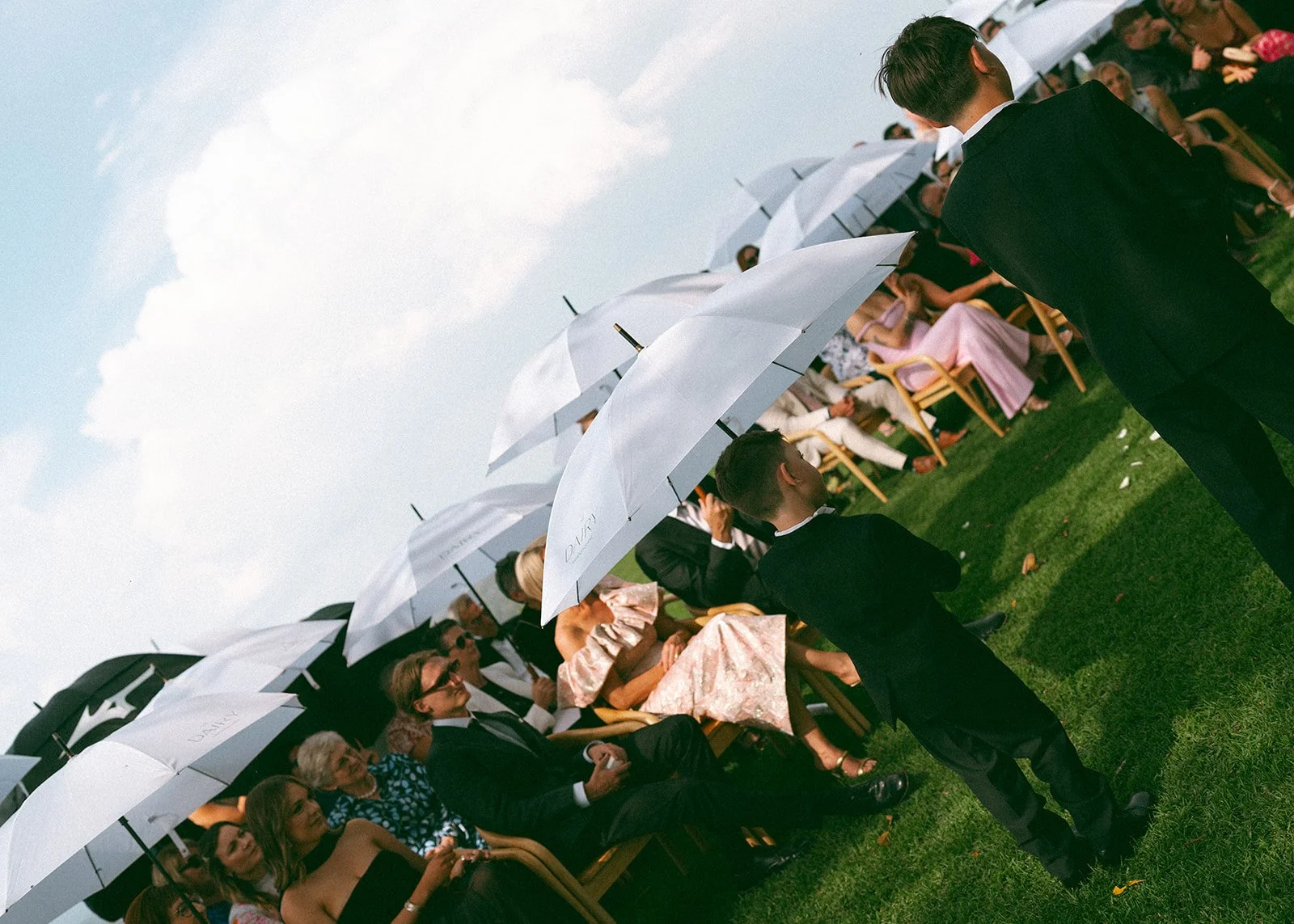 People attending an outdoor event on a lawn, seated under white umbrellas, dressed in formal attire, with some standing and some sitting, with a mostly cloudy sky overhead.