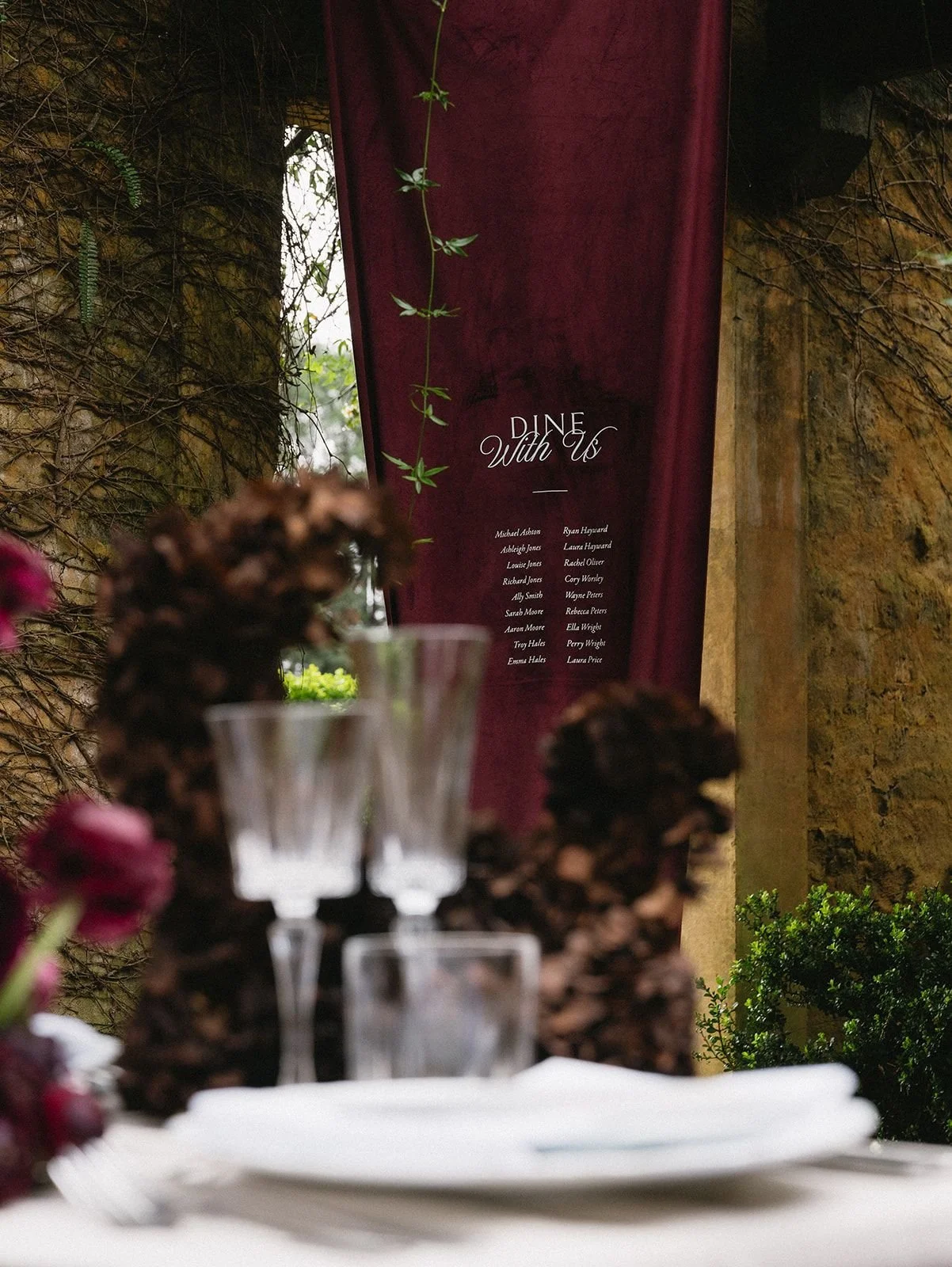 A formal dining setup with wine glasses, napkins, and floral centerpieces, with a burgundy curtain and a menu board in the background.
