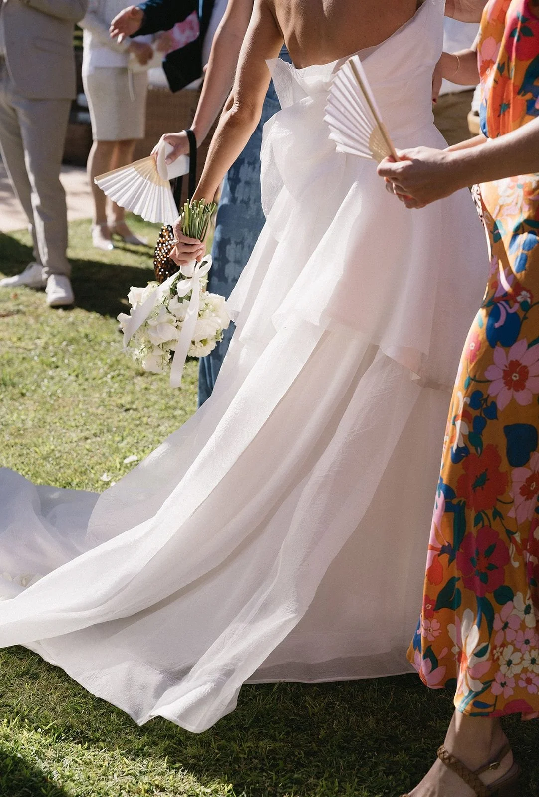 Close-up of a bride wearing a white wedding gown holding a bouquet of white flowers and a fan, standing on grass during an outdoor wedding ceremony. Guests in colorful and neutral outfits are visible in the background.