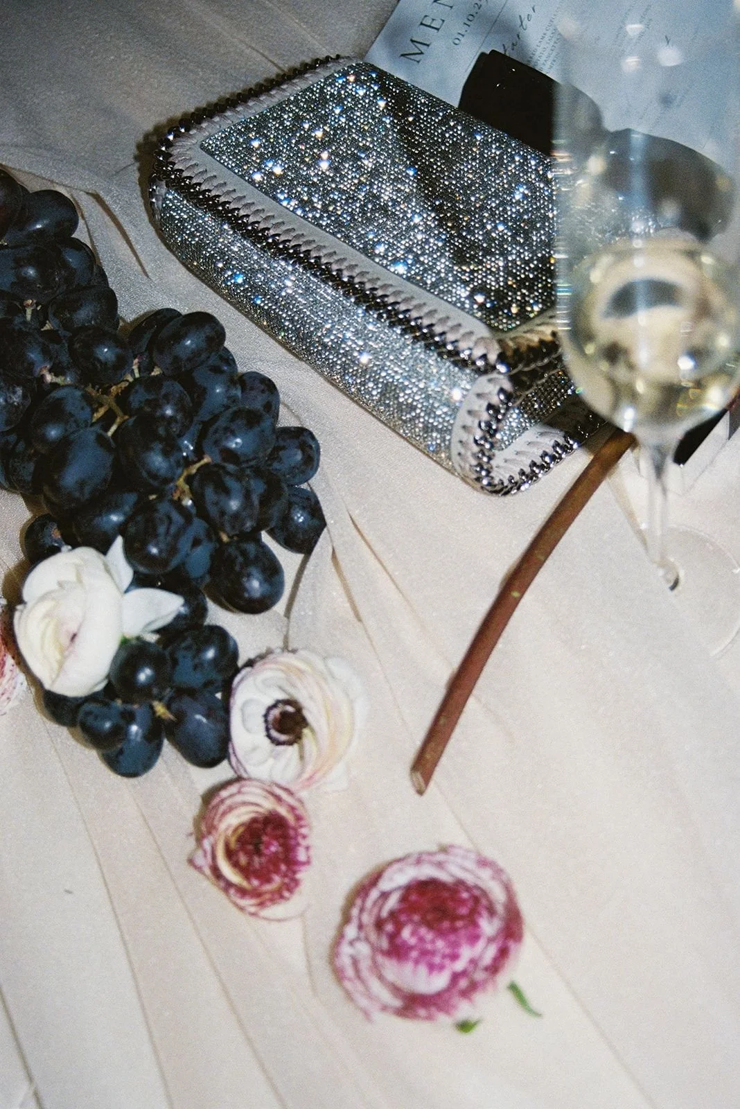 A glittery, rhinestone-encrusted clutch purse, a bunch of dark grapes, and a partially visible glass of white wine on a beige tablecloth with scattered flower petals and a newspaper.