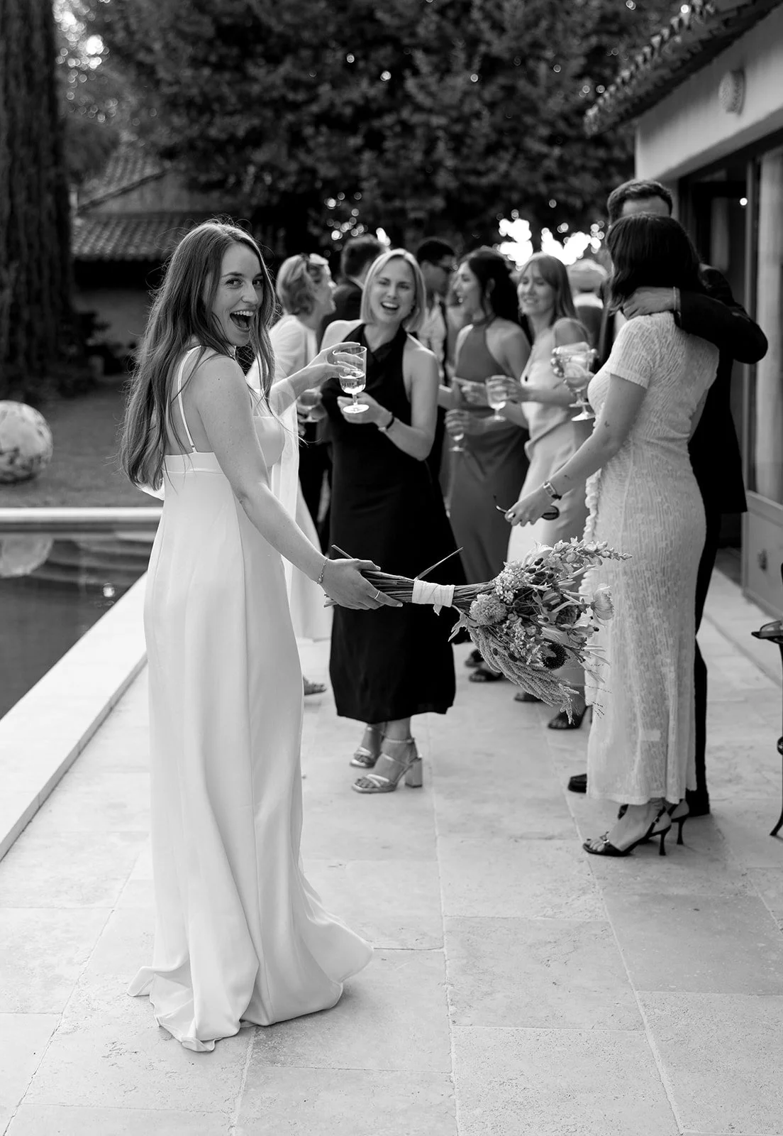A woman in a white dress holding a bouquet of flowers, smiling and holding a glass, at a gathering of people near a pool, with others holding drinks and talking.