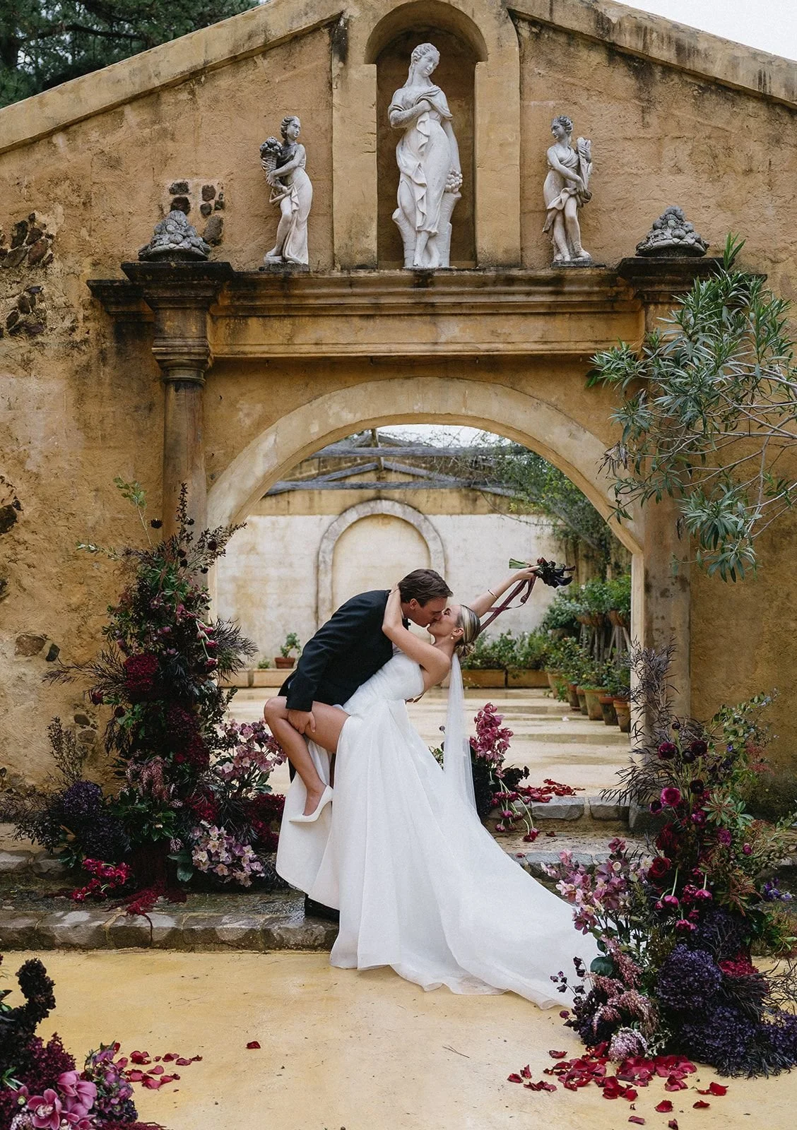 A wedding couple is kissing under an ancient stone archway, surrounded by flower arrangements, with a decorative wall featuring classical statues above.