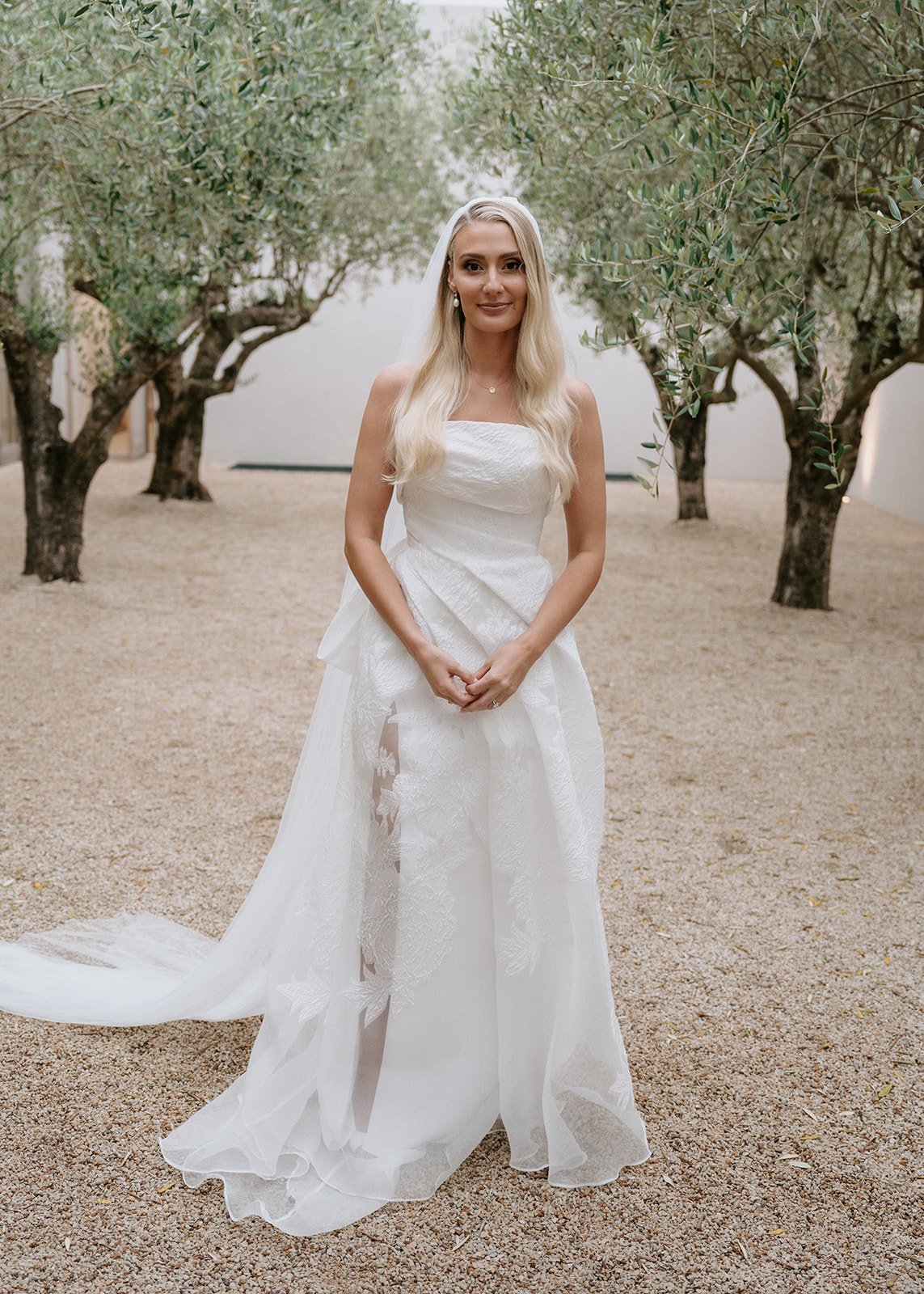 A woman in a white wedding dress standing in an outdoor setting with trees and a gravel ground.