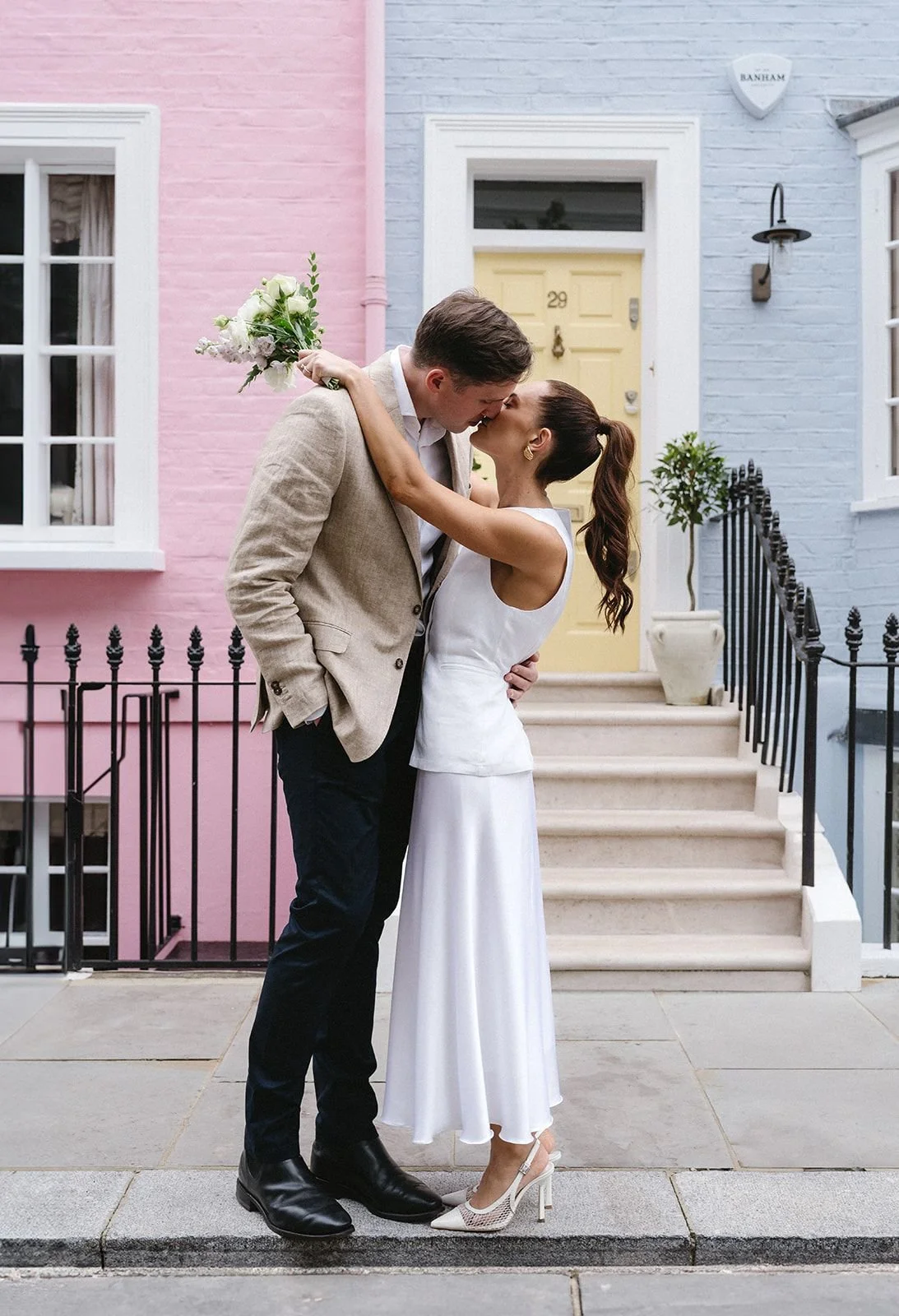 A couple kissing on steps outside a pastel-colored house, woman in white dress and high heels, man in beige blazer and black shoes, woman holding a bouquet of flowers.
