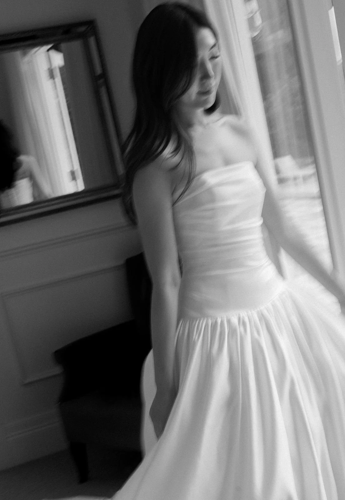A woman in a strapless gown standing by a window, looking down, in black and white.