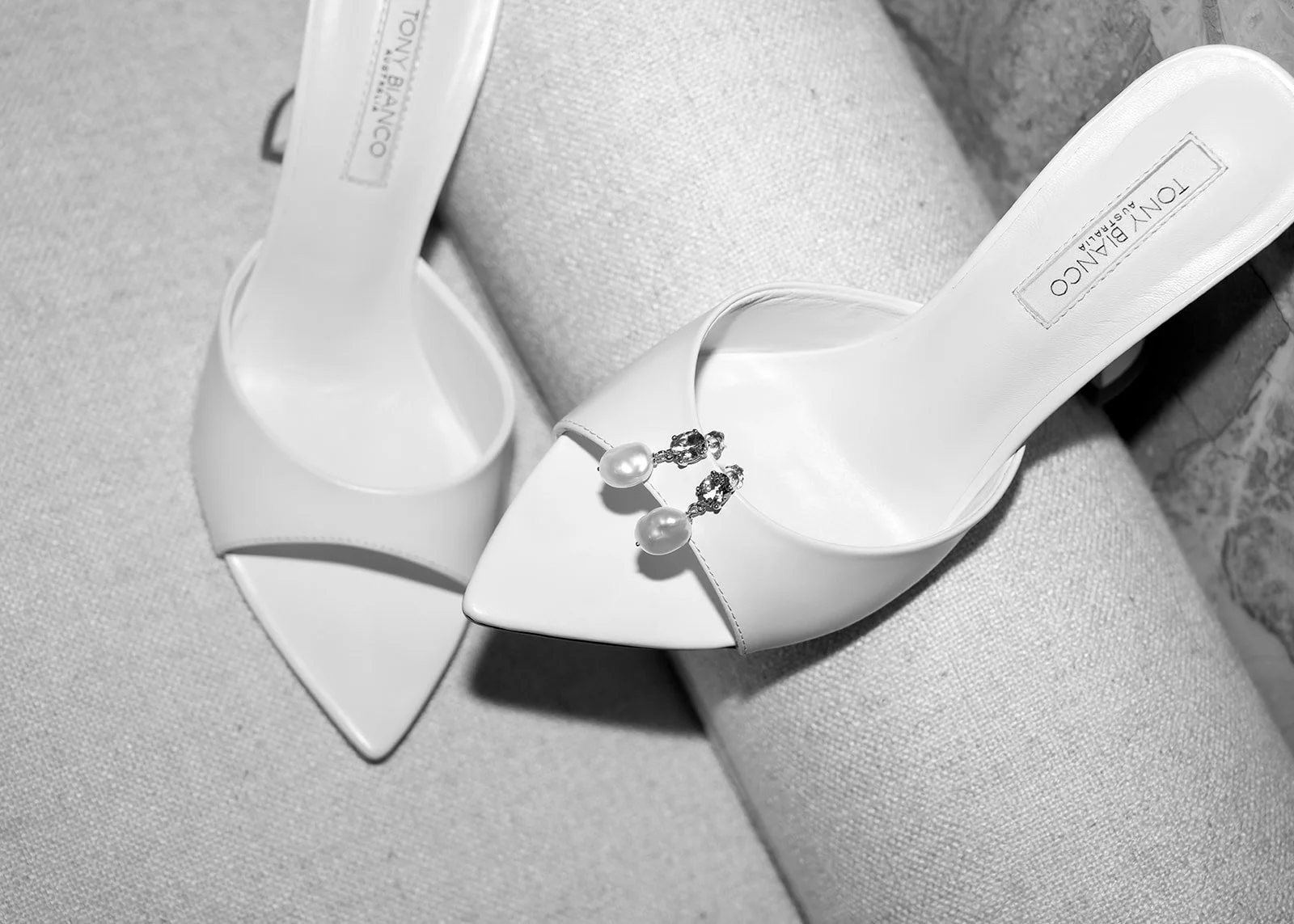 A pair of white high-heeled shoes with decorative pearl and rhinestone embellishments on the front, placed on a textured light-colored surface.