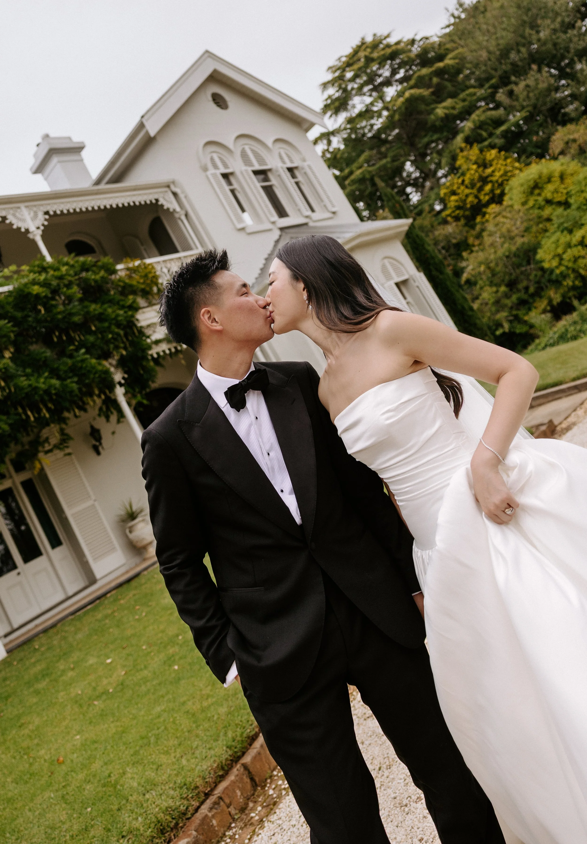 Brontë McVeigh wedding photography: A newlywed couple sharing a kiss in front of a large white Victorian-style house with trees and greenery in the background.