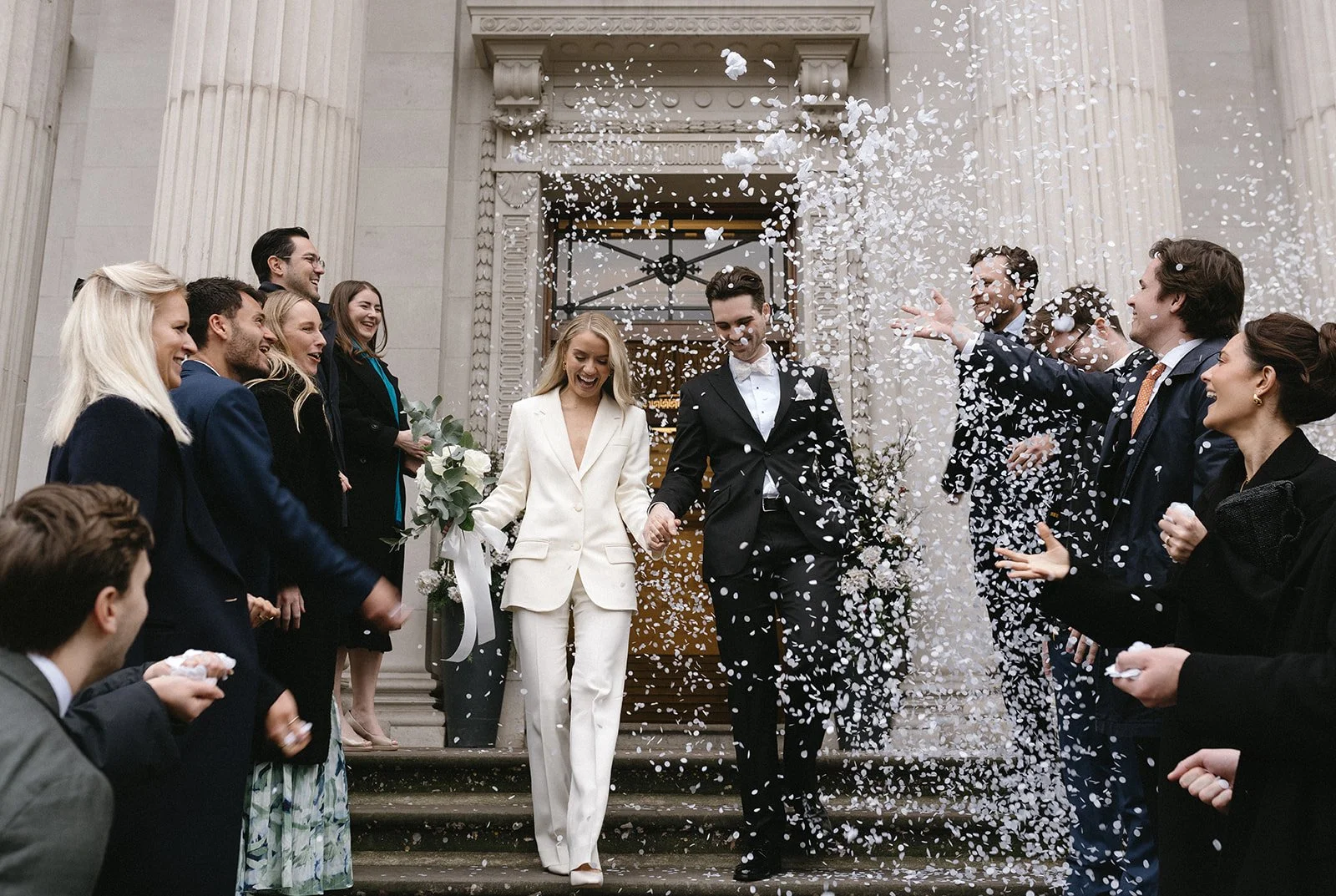 Wedding photography by Brontë McVeigh: A newlywed couple celebrating on the steps of a historic building as friends and family throw confetti in the air.
