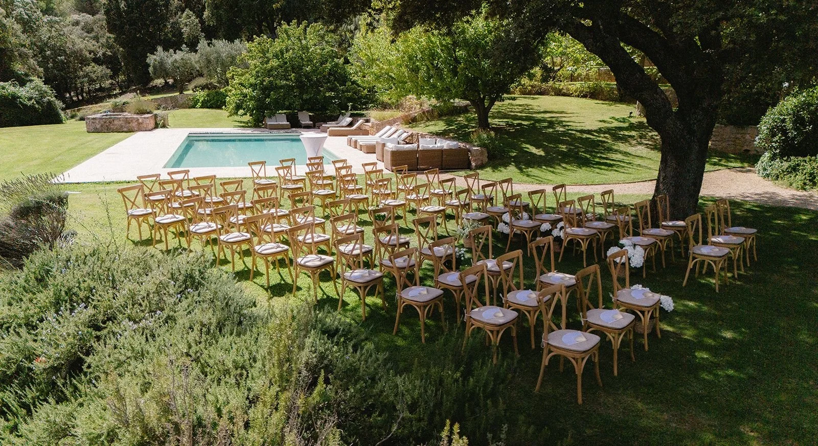 Outdoor wedding setup with rows of wooden chairs on a grassy lawn, facing a pool, shaded by large trees, with a seating area and greenery in the background.