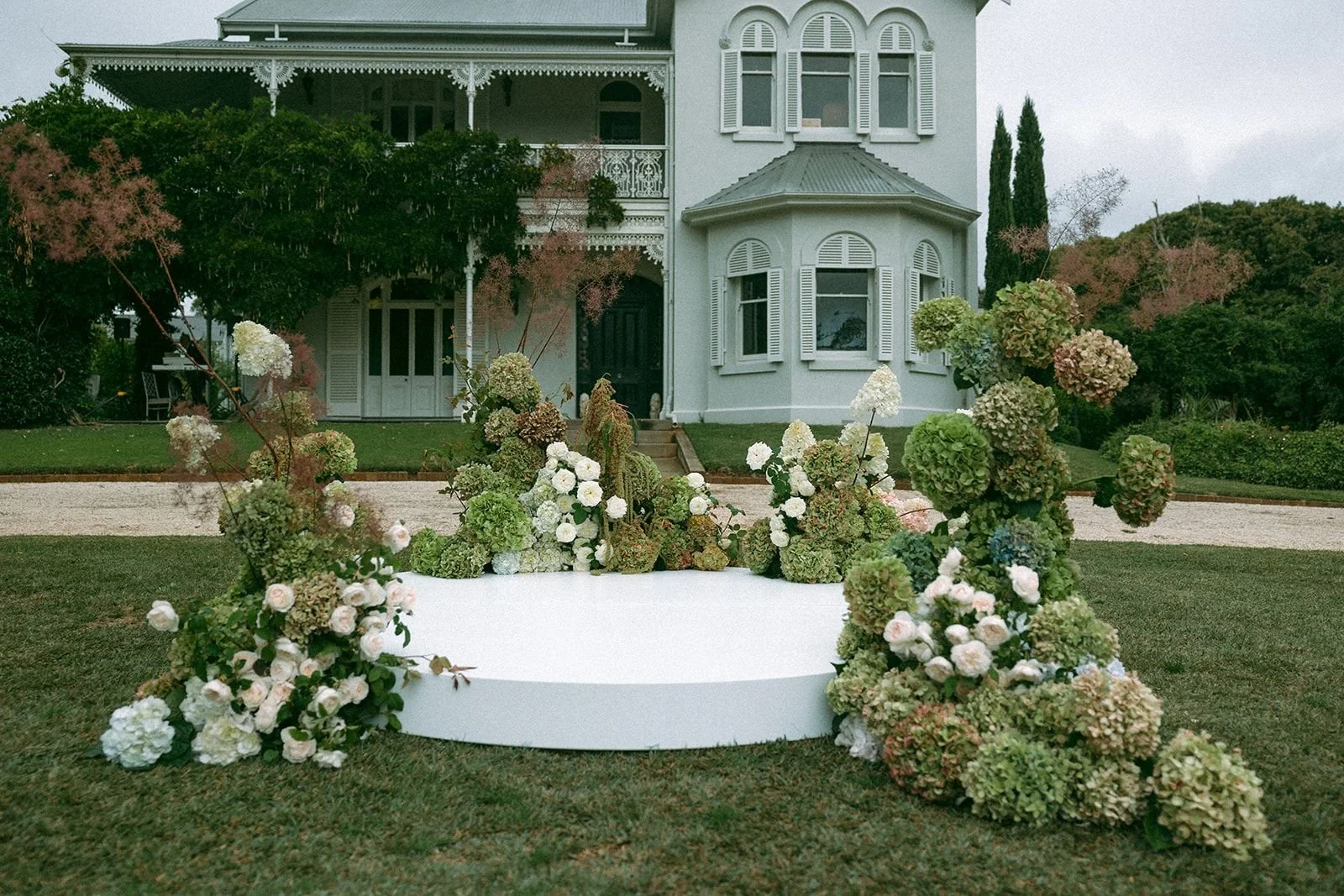 Outdoor scene with a white Victorian-style house in the background. In the foreground, there is a white circular platform surrounded by a large arrangement of green and white hydrangeas and other flowers, possibly set up for an outdoor event or weddi