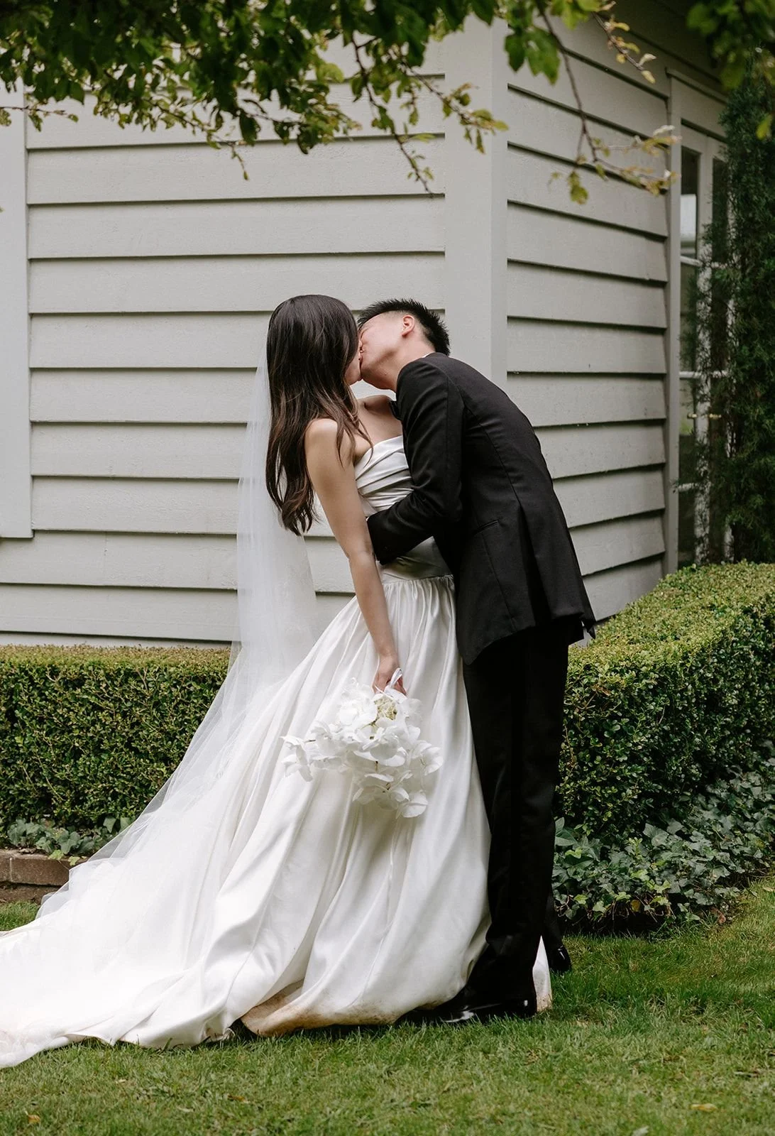 A wedding couple sharing a kiss outdoors, the bride in a white wedding gown holding a bouquet of white flowers, the groom in a black suit, standing on a grassy area near bushes and a gray house.