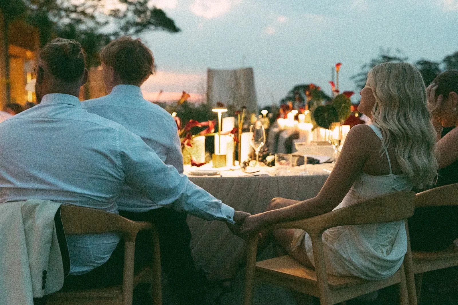 A couple holding hands at an outdoor dinner gathering during sunset, surrounded by other guests and decorated with flowers and candles.