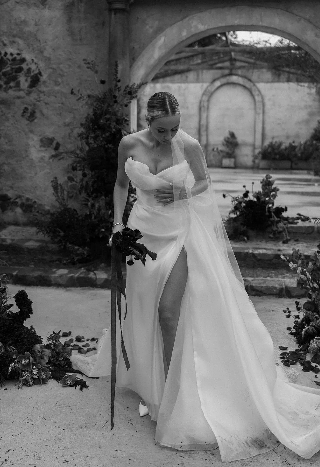 A bride in a wedding dress holding a bouquet, standing outdoors in a stone courtyard with arches and plants, in black and white.