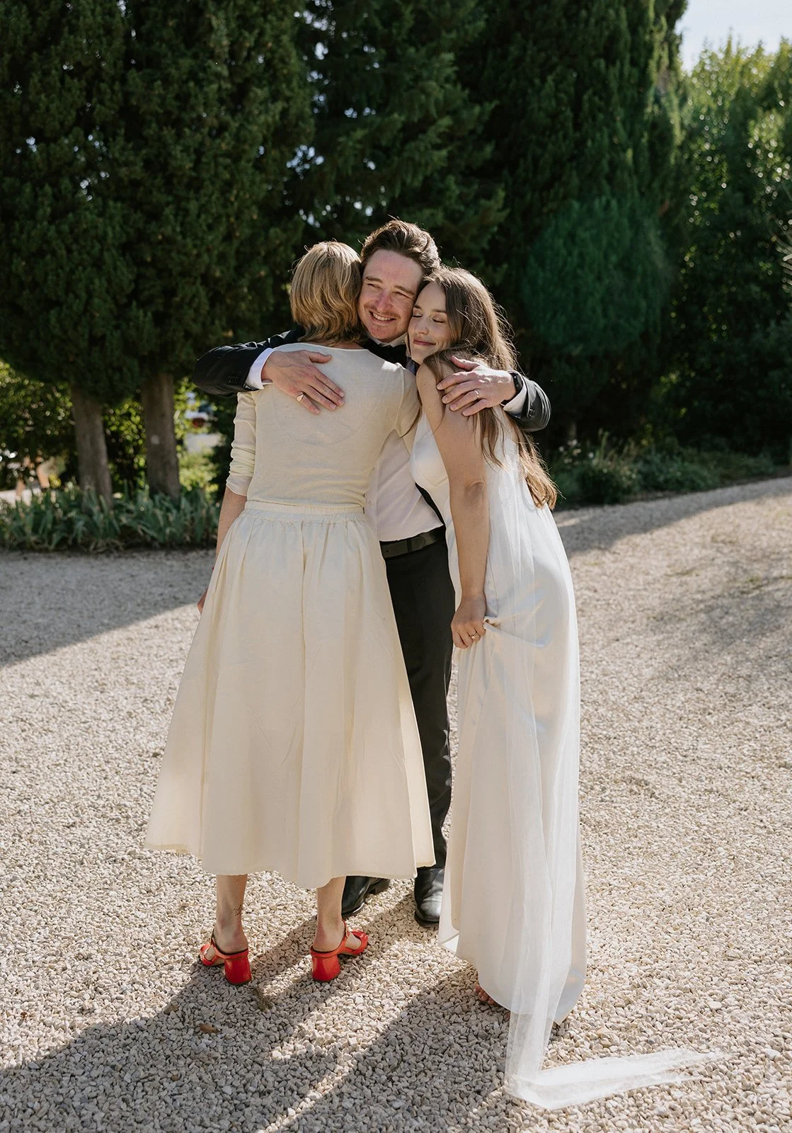 Three people embracing outdoors on a sunny day, with lush green trees in the background, two women in white dresses and a man in black trousers and a white shirt.