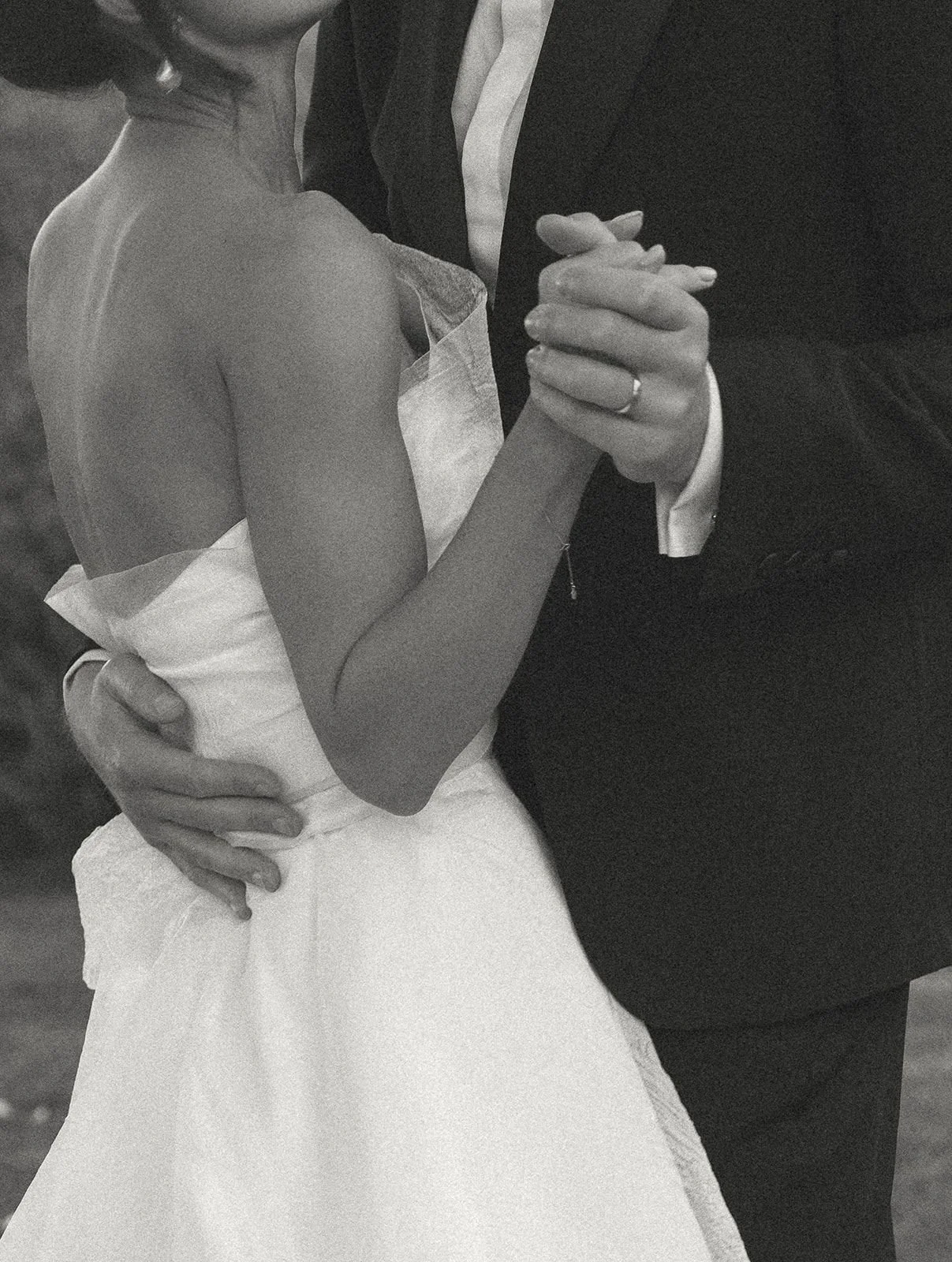 A black-and-white photo of a couple dancing closely, holding hands with intertwined fingers, the woman's hand resting on the man's shoulder, the woman's wedding dress and the man's suit are partially visible.