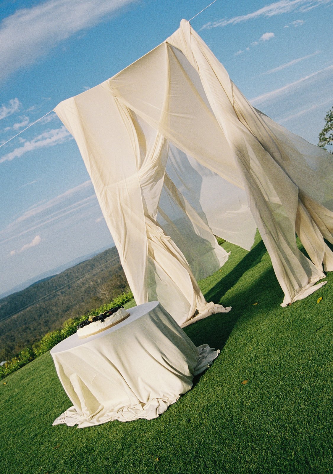 Outdoor setup with a fabric tent, a table with a cake, on green grass with hills and a blue sky in the background.