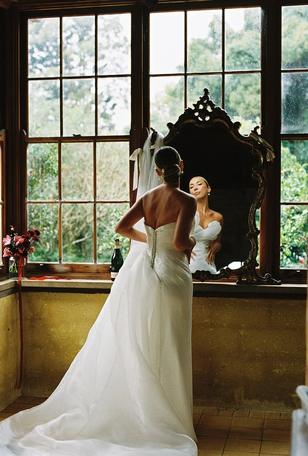 Bride in wedding dress looking at herself in a large ornate mirror in a room with big windows and greenery outside.