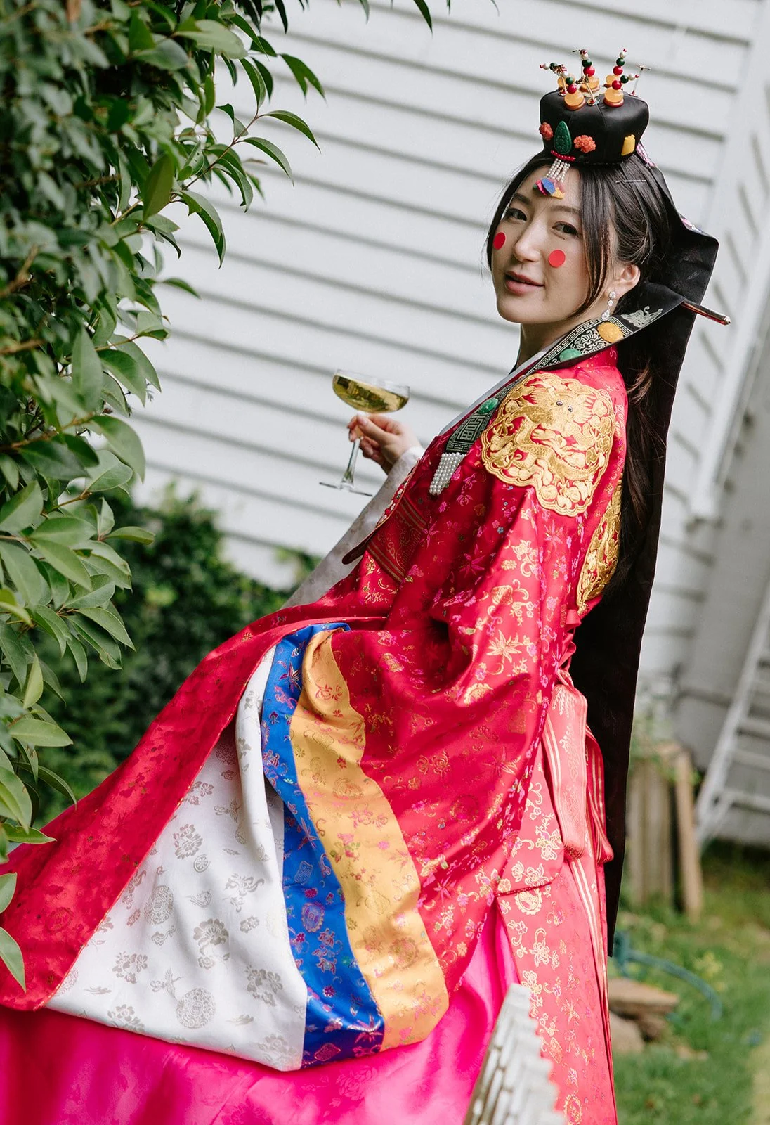 Woman wearing traditional Korean hanbok with elaborate headpiece, holding a glass of champagne, standing outdoors near greenery and a white house.