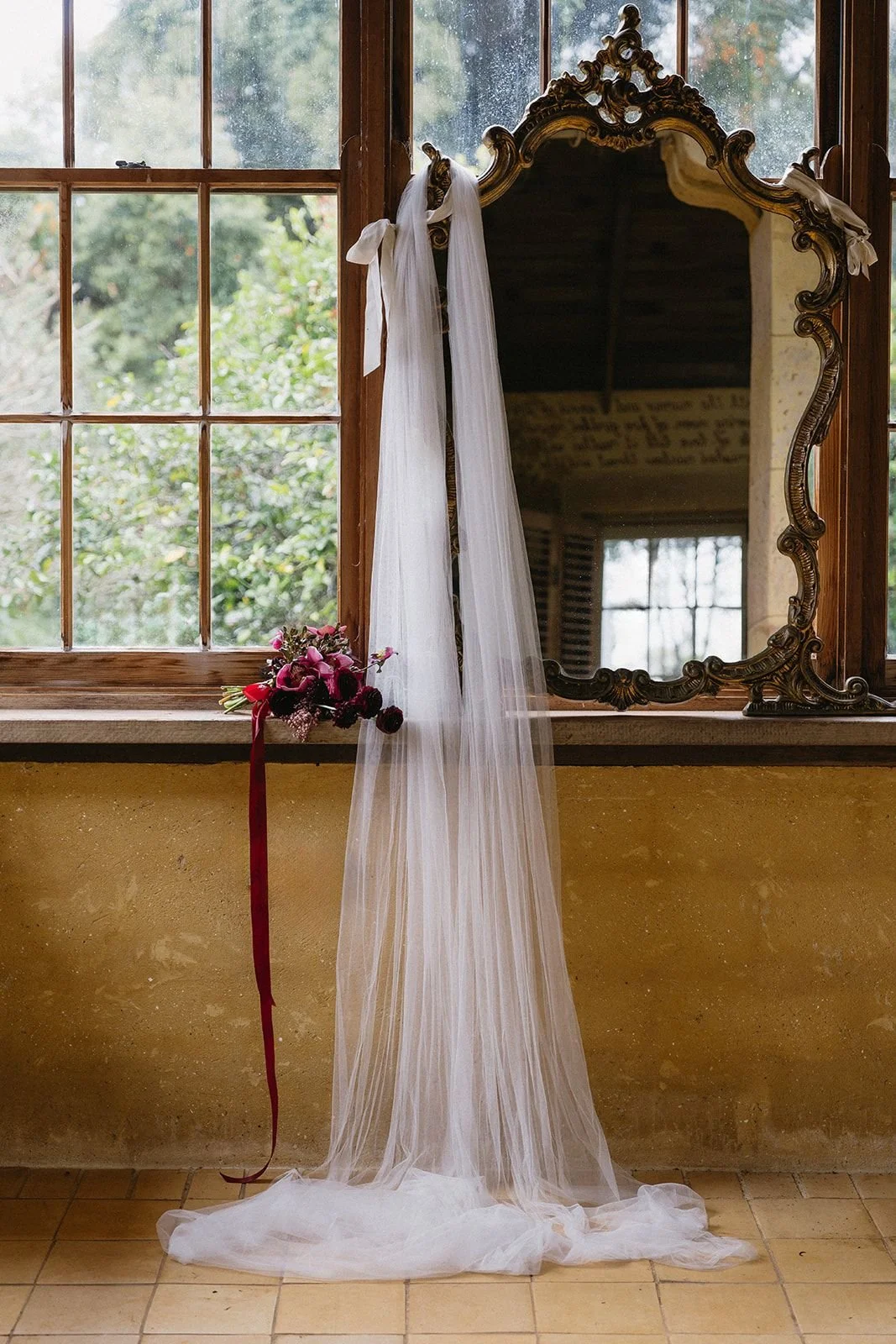 Wedding veil hanging on a window with a bouquet of flowers resting on the window sill, a large ornate mirror, and a window showing greenery outside.