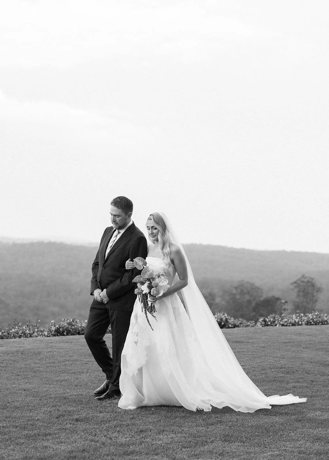 A bride and groom walking together outdoors on a grassy field, holding hands, with a landscape of hills and trees in the background, black and white photograph.
