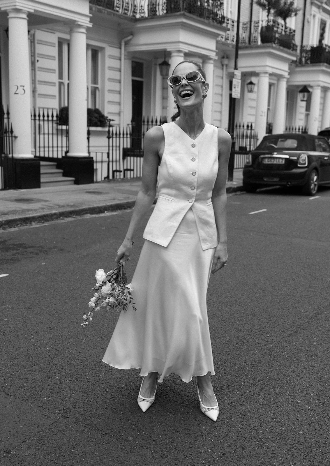 A woman in a white dress and large sunglasses is smiling and holding a bouquet of flowers on a city street.