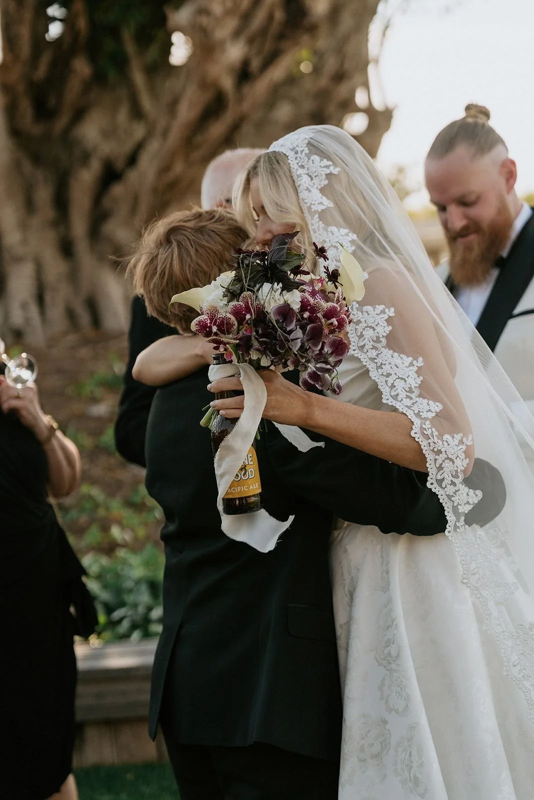 A bride in a wedding dress and veil hugs a person holding a bouquet of flowers during a wedding ceremony, with other guests in the background.