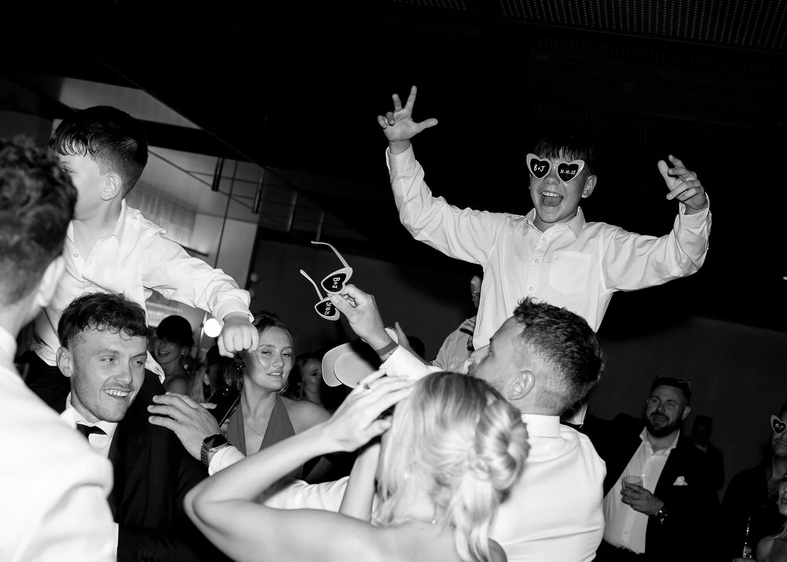 People celebrating at a wedding reception, with two young boys on their shoulders, wearing heart-shaped sunglasses, and holding heart-shaped sunglasses as they dance.
