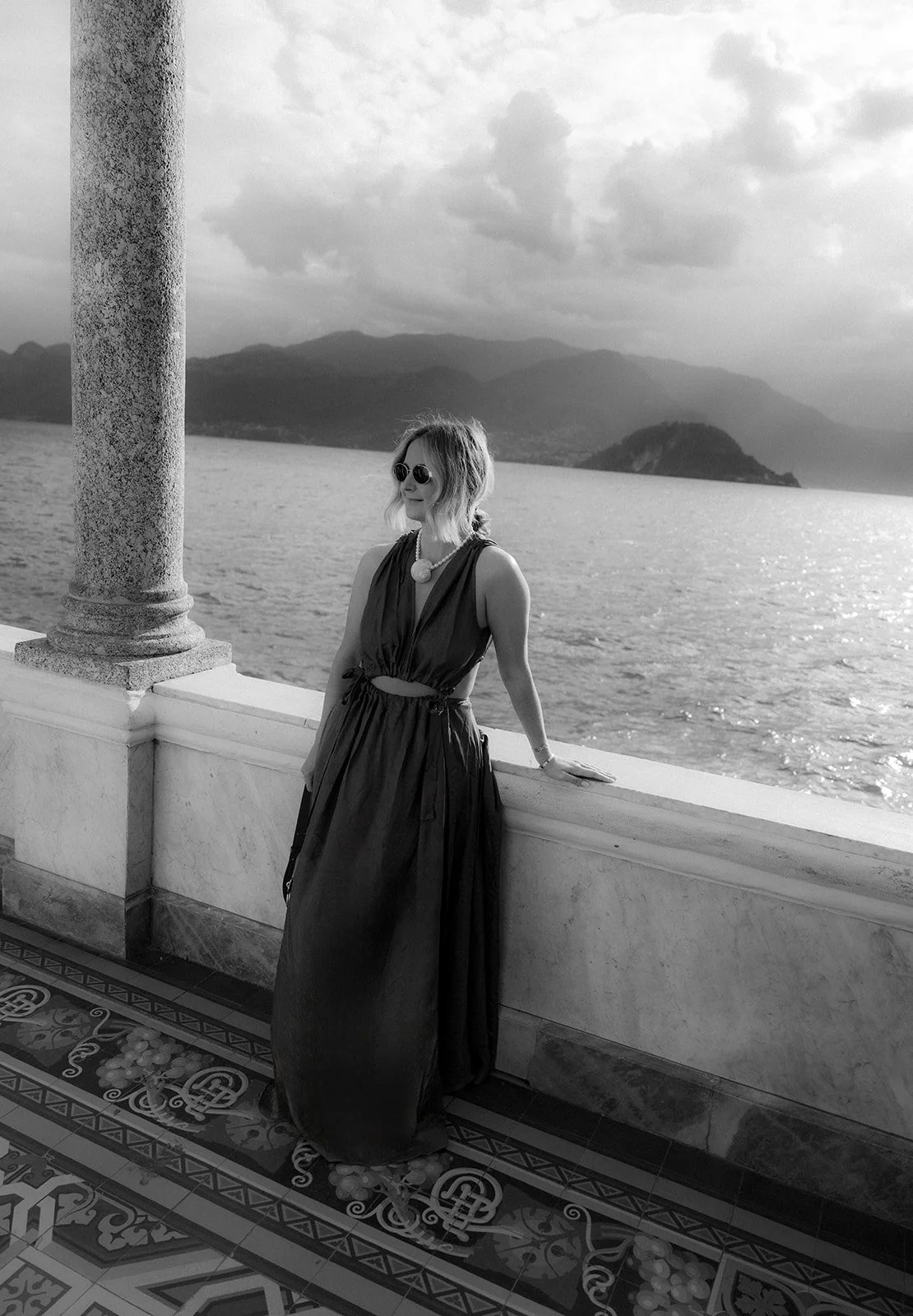 Brontë McVeigh: A woman wearing sunglasses and a sleeveless dress stands beside a balustrade overlooking a body of water with mountains in the background, in a black-and-white photo.