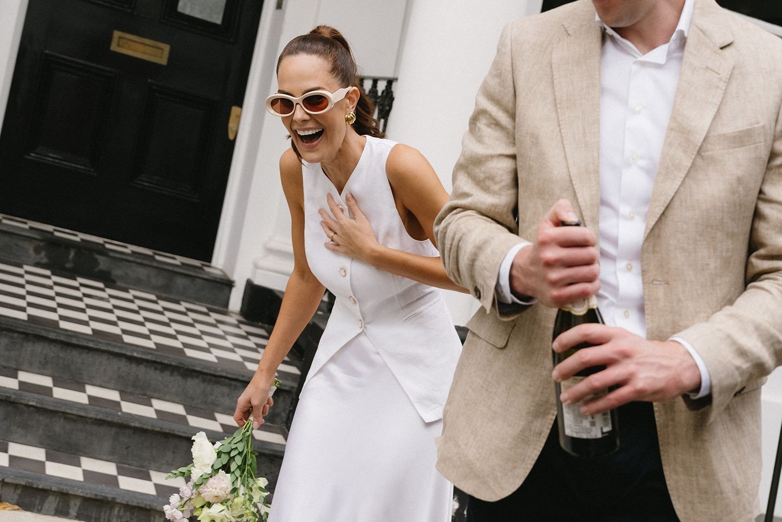 A woman in a white dress and sunglasses, laughing and holding a bouquet, stands on a staircase with a man in a beige suit holding a beer bottle, in an indoor setting.