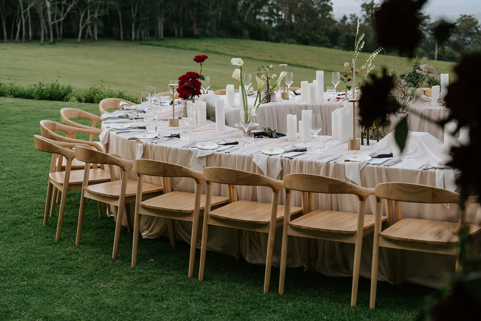 Elegant outdoor dining table set on grass with wooden chairs, white tablecloth, and floral centerpieces, surrounded by greenery.
