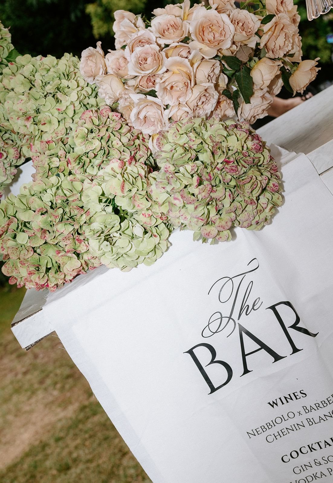 Bouquets of pale pink roses and white-pink hydrangeas with a sign reading "The Bar" offering wines, cocktails, and other beverages.