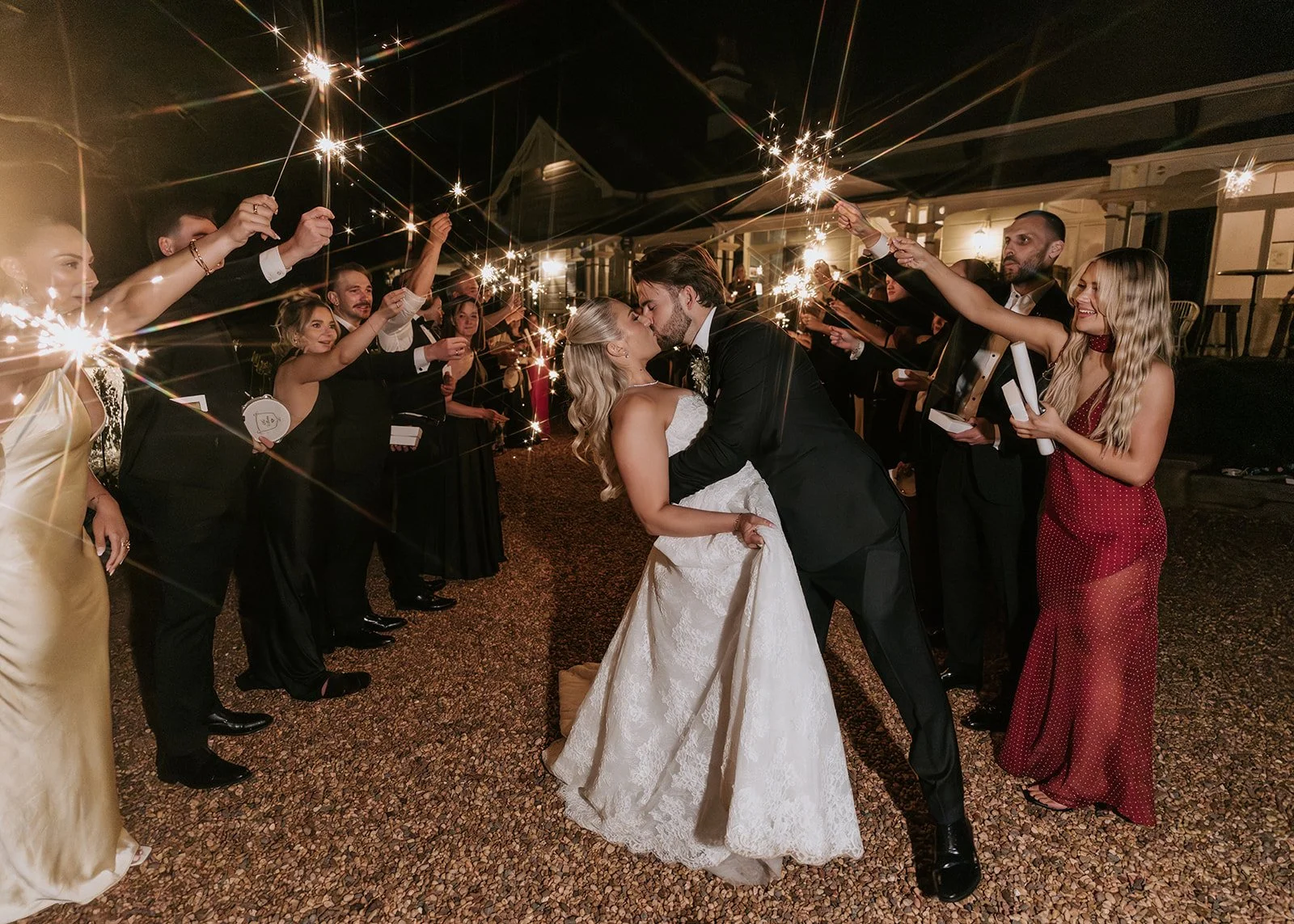 Bride and groom kiss while surrounded by guests holding sparklers at night during wedding celebration.