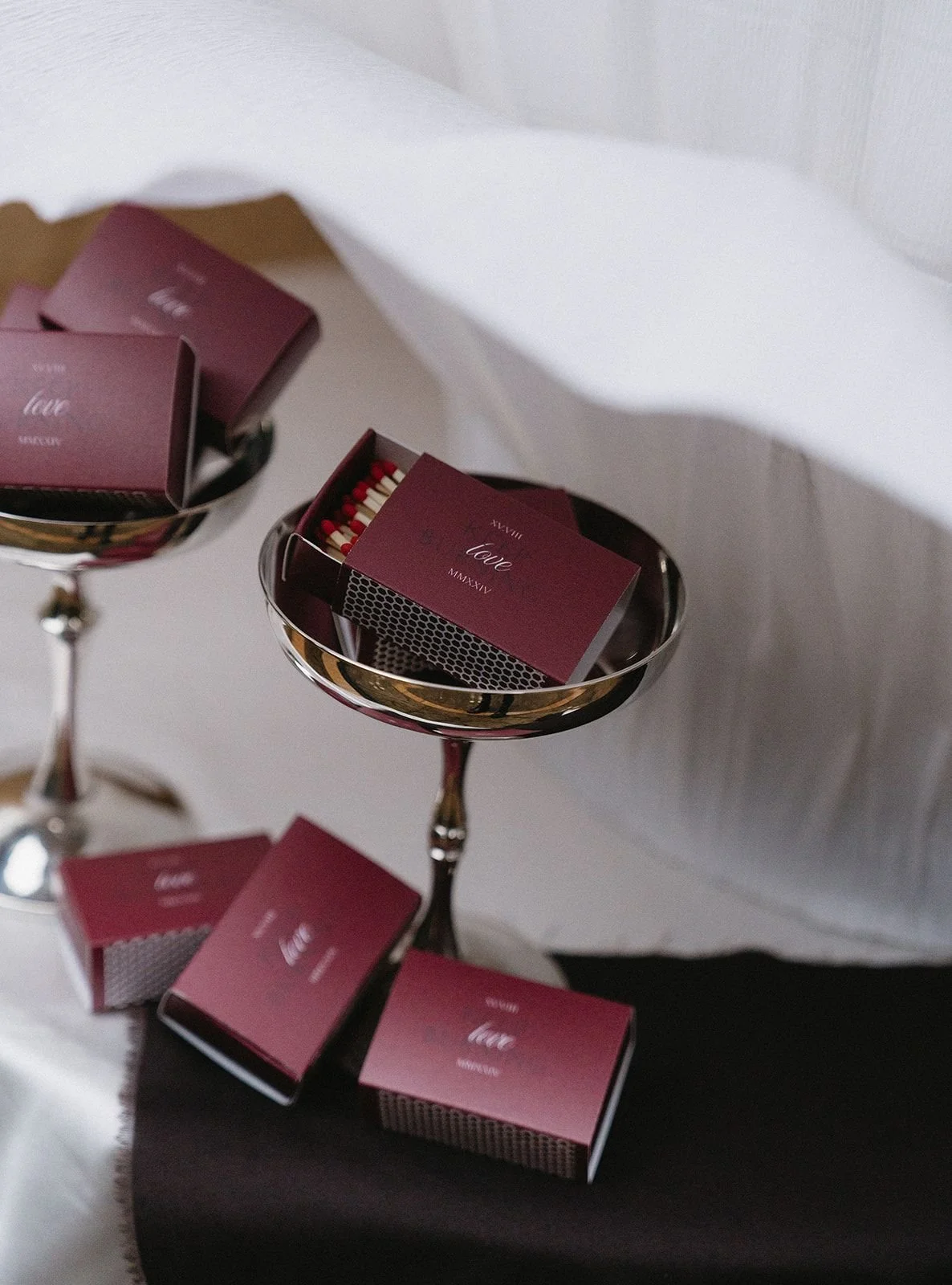 Two silver trays holding small boxes of matches with maroon packaging on a dark oval table with a white textured wall background.