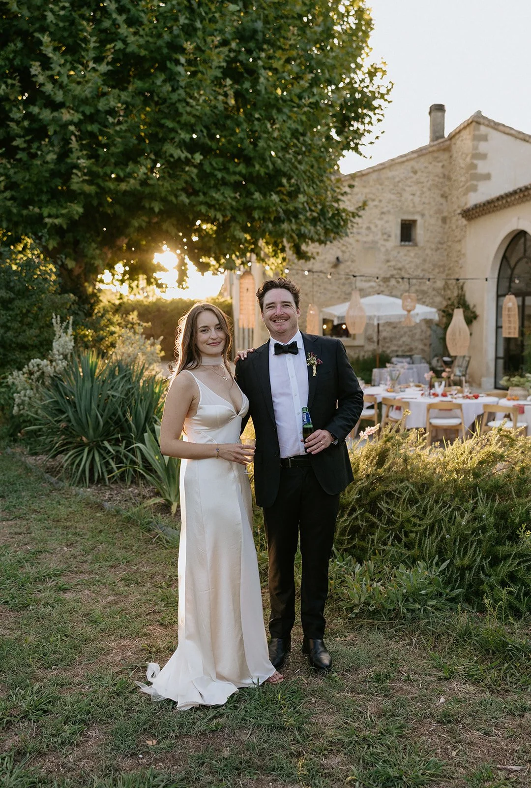 A man and woman in formal attire smiling at a wedding reception outdoors during sunset. The woman is in a white gown holding a drink, and the man is in a tuxedo holding a bottle. There are tables set up with decorations in the background, surrounded 