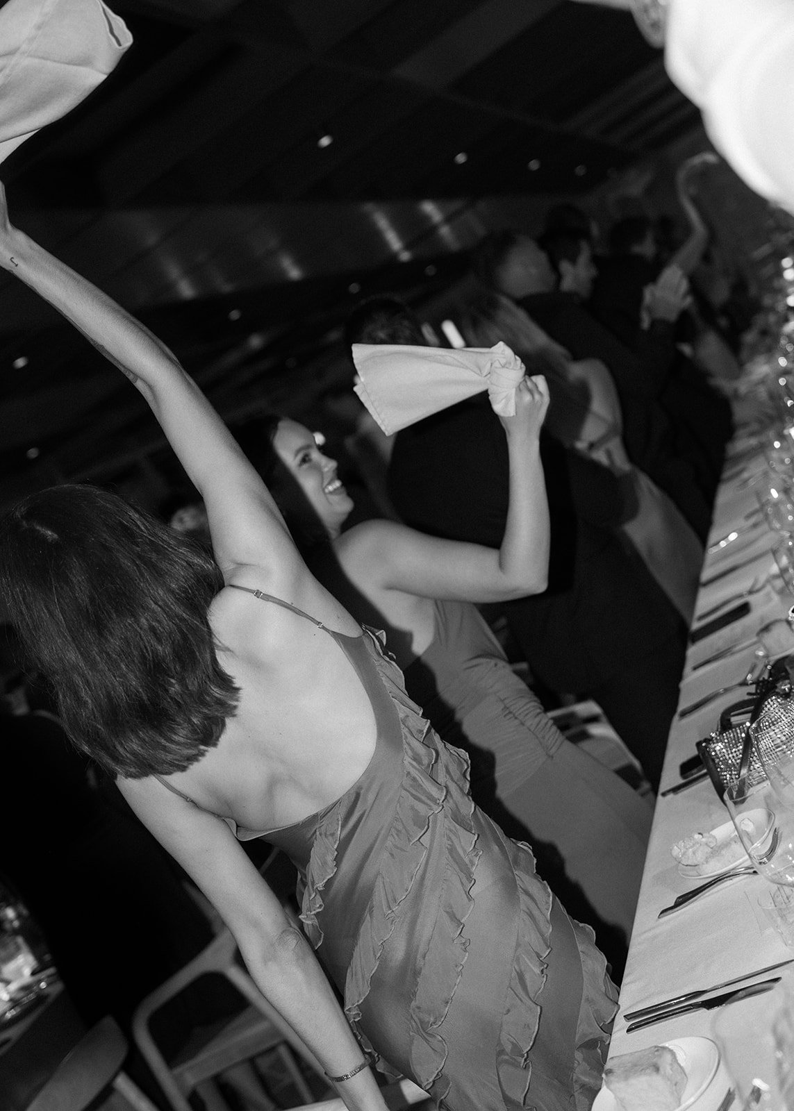 People sitting at a formal event or dinner, with some smiling and raising their hands.