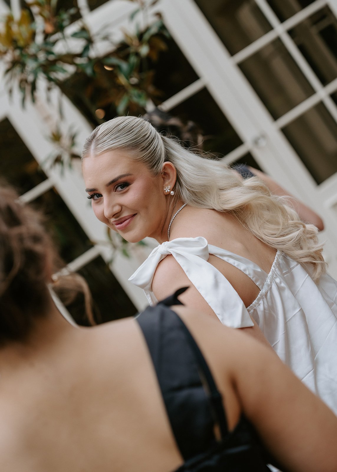 A smiling blonde woman in a white dress with ribbons on the shoulders, wearing pearl earrings and a necklace, looking over her shoulder at someone else during a social gathering.